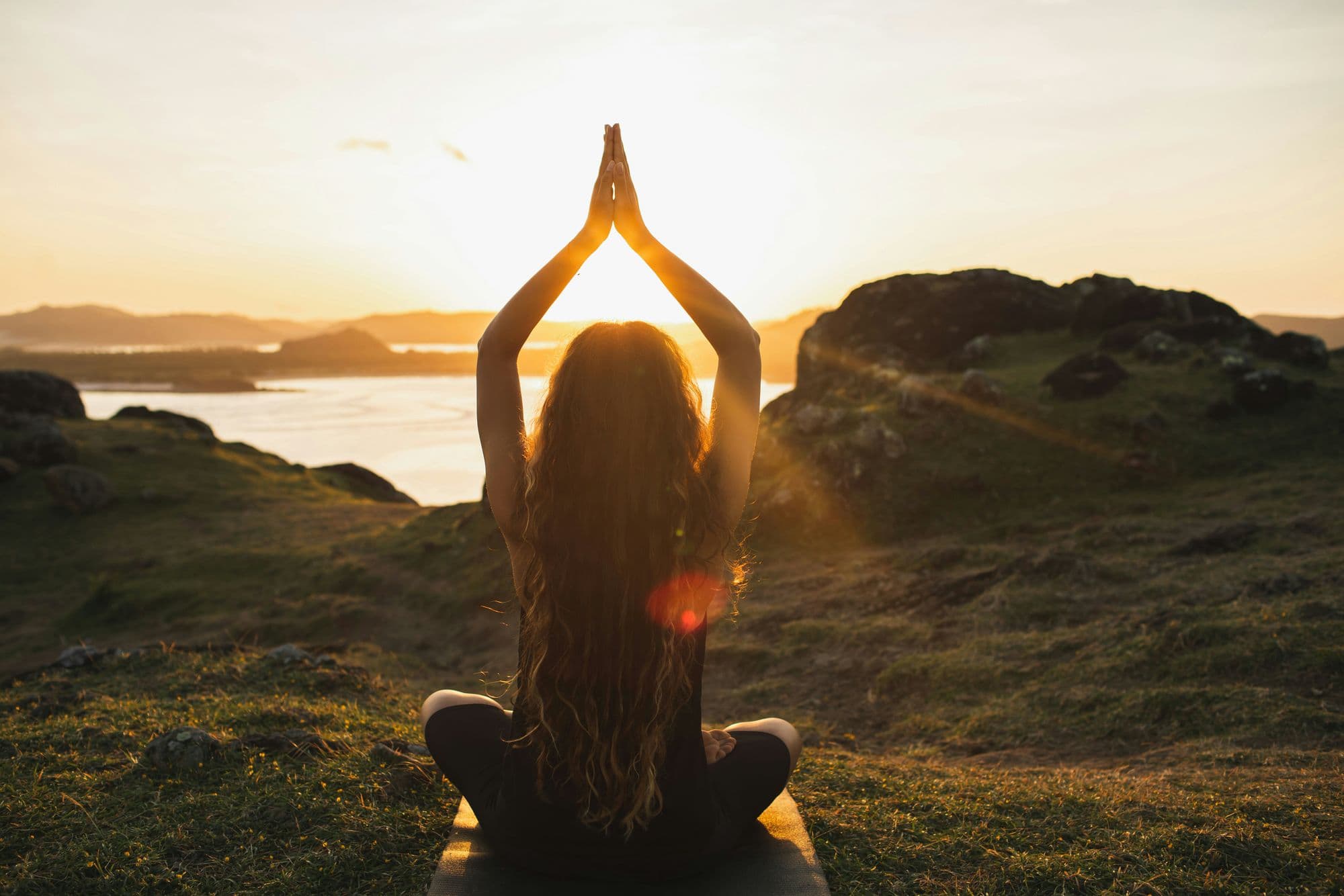 Woman meditating cross-legged at sunset with hands raised overhead, overlooking the sea